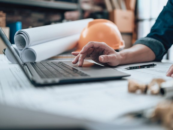 Hand am Laptop auf Bauzeichnungen, im Hintergrund ein Bauhelm und Pläne