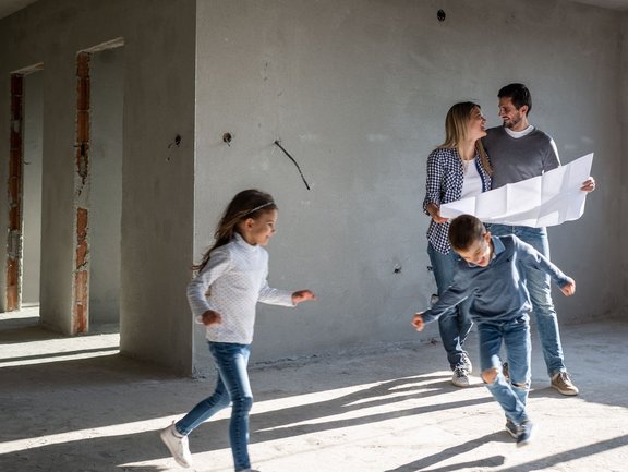 Eltern mit Bauplan und zwei spielenden Kindern im Rohbau eines Hauses