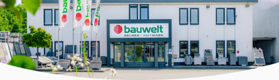 Außenansicht des Hauptgebäudes der Firma "bauwelt Delmes Heitmann" am Standort Hummelsbüttel mit weißer Metallfassade, modernem Eingangsbereich und großem Firmenschild. Vor dem Gebäude befinden sich mehrere bauwelt-Flaggen sowie Ausstellungsflächen mit Boden- und Wandbelägen. Links und rechts sind weitere Materialien und Musterflächen zu sehen, eingerahmt von Bäumen und Begrünung.