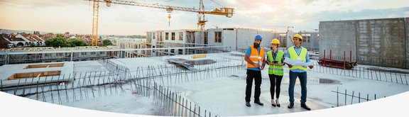 Drei Bauarbeiter stehen auf einer Baustelle, im Hintergrund Kräne und Gebäude