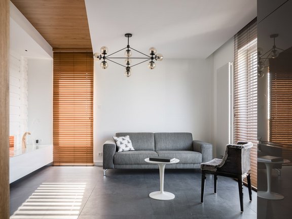 Minimalistisches Wohnzimmer mit grauem Sofa, modernen Jalousien und weißen Wänden, Holzdetails an Decke und Fensterrahmen