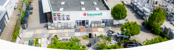 ußenansicht des Hauptgebäudes der Firma "bauwelt Delmes Heitmann" am Standort Kaltenkirchen mit heller Metallfassade und großem Logo über dem Eingangsbereich. Vor dem Gebäude befinden sich großzügige Ausstellungsflächen mit verschiedenen Pflastersteinen, Fliesen und Gestaltungselementen. Rechts ist ein Lagerbereich mit Regalsystemen und Baustoffen sichtbar, eingerahmt von Bäumen und Grünflächen.