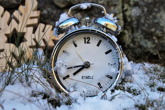 Alter silberner Wecker liegt von Schnee bedeckt im Gras, Zeiger zeigen auf 7:45 Uhr