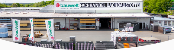 Außenansicht des Standorts der Firma "bauwelt Delmes Heitmann" in Harburg mit Fokus auf den Fachhandel für Dachbaustoffe. Das Gebäude hat eine breite Lagerhalle mit offenem Wareneingang, Regalen mit Baumaterialien und mehreren großen Firmenschildern. Im Vordergrund befinden sich Verkaufs- und Ausstellungsflächen für Dachziegel sowie mehrere „bauwelt“-Flaggen.