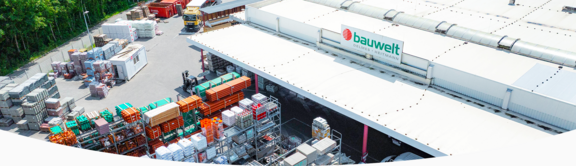 Außenansicht des Standorts der Firma "bauwelt Delmes Heitmann" in Winsen mit großflächiger Lagerhalle und überdachtem Warenbereich. Auf dem Hof sind zahlreiche Baustoffe wie Rohre, Paletten, Steine und Baumaterialien aufgestapelt. Ein Gabelstapler ist im Einsatz, im Hintergrund sind LKWs und Container zu sehen. Das Gelände ist von Bäumen umgeben.