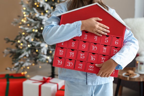 Kind im Schlafanzug hält einen roten Adventskalender in Form eines Hauses vor einem Weihnachtsbaum