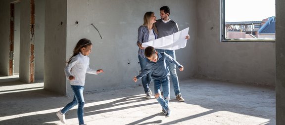 Eltern mit Bauplan und zwei spielenden Kindern im Rohbau eines Hauses