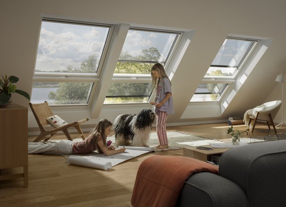 Helles Dachgeschosszimmer mit großen Dachfenstern, zwei spielenden Kindern und einem Hund auf einem Teppichboden, gemütlich eingerichtet mit Holz- und Stoffmöbeln.