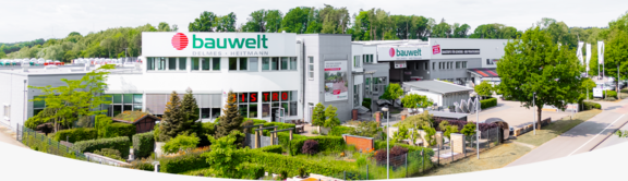 Außenansicht des Hauptgebäudes der Firma "bauwelt Delmes Heitmann" am Standort Seevetal mit modernem, weiß-silbernem Gebäudekomplex, großem Logo auf der Fassade und einem gepflegten Außenbereich mit Garten- und Landschaftselementen. Rechts verläuft eine Straße mit Bäumen, im Hintergrund sind weitere Gebäude und ein Waldstück zu sehen.