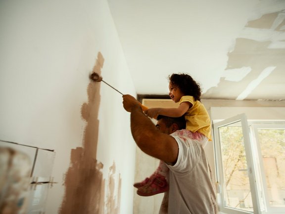Vater trägt seine Tochter auf den Schultern, während sie gemeinsam eine Wand mit Farbe streichen
