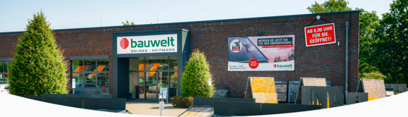 Außenansicht des Hauptgebäudes der Firma "bauwelt Delmes Heitmann" am Standort Hollenstedt mit modernem Backsteingebäude, großem Eingangsbereich und prominentem Firmenschild. An der Fassade hängt ein Werbebanner zur Energiewende sowie ein Schild mit der Aufschrift „Ab 6.00 Uhr für Sie geöffnet!“. Im Vordergrund befinden sich Ausstellungen mit Dach- und Fassadenmaterialien, eingerahmt von gepflegten Pflanzen.