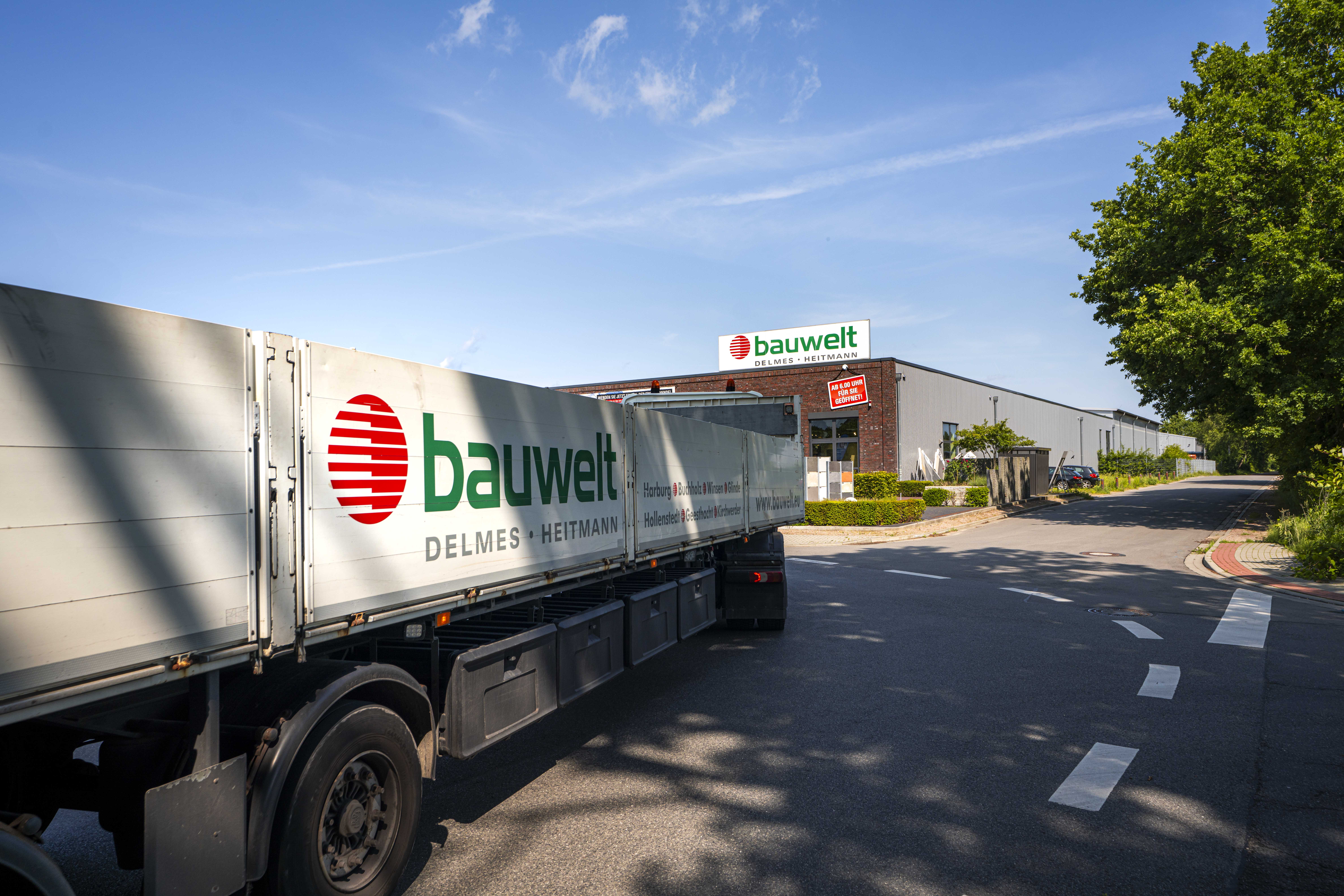 Ein Bauwelt-LKW mit großem Logo fährt auf eine Straße vor dem Bauwelt-Standort Hollenstedt zu, dessen Gebäude mit Klinkerfassade, Firmenlogo und Werbetafel im Hintergrund zu sehen ist. Der Himmel ist blau und die Umgebung grün bepflanzt.