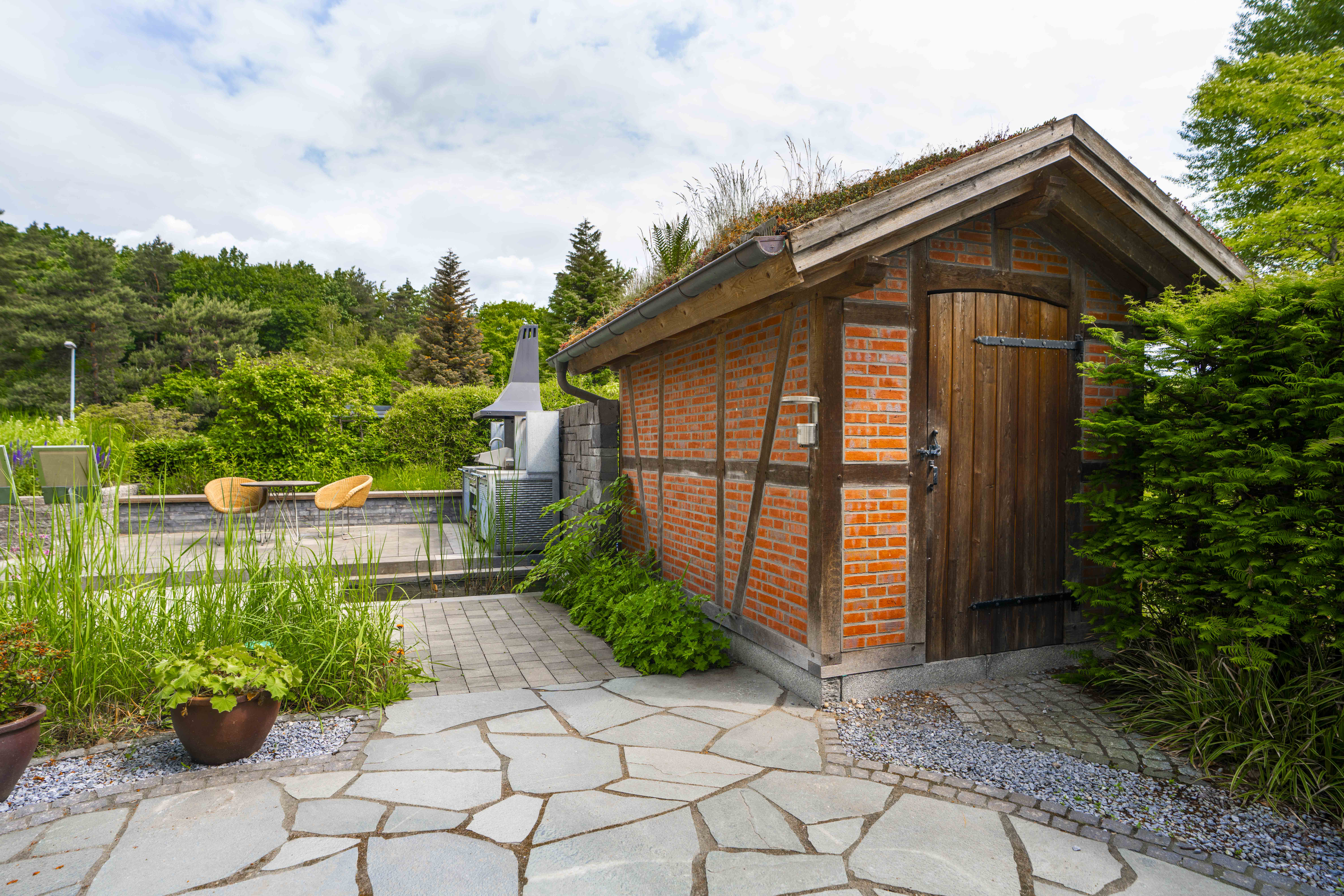 Kleines Gartenhaus aus Backstein mit Holzdetails neben einer Terrasse mit Gartenmöbeln und Grillplatz.