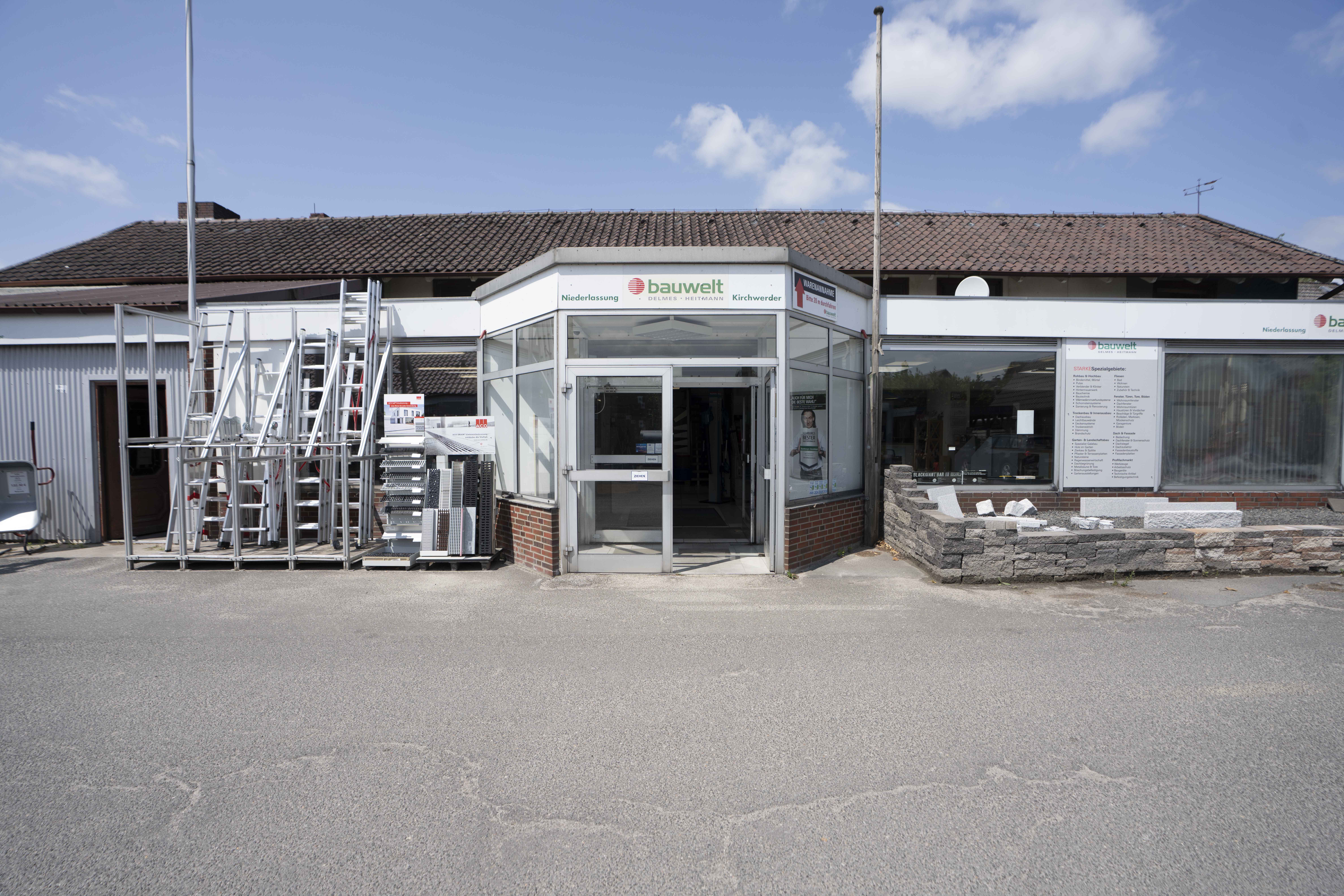 Frontalansicht des Eingangsbereichs vom Bauwelt-Standort Kirchwerder mit Glasfassade, Außendisplay für Bauelemente und angrenzendem Fachmarkt.