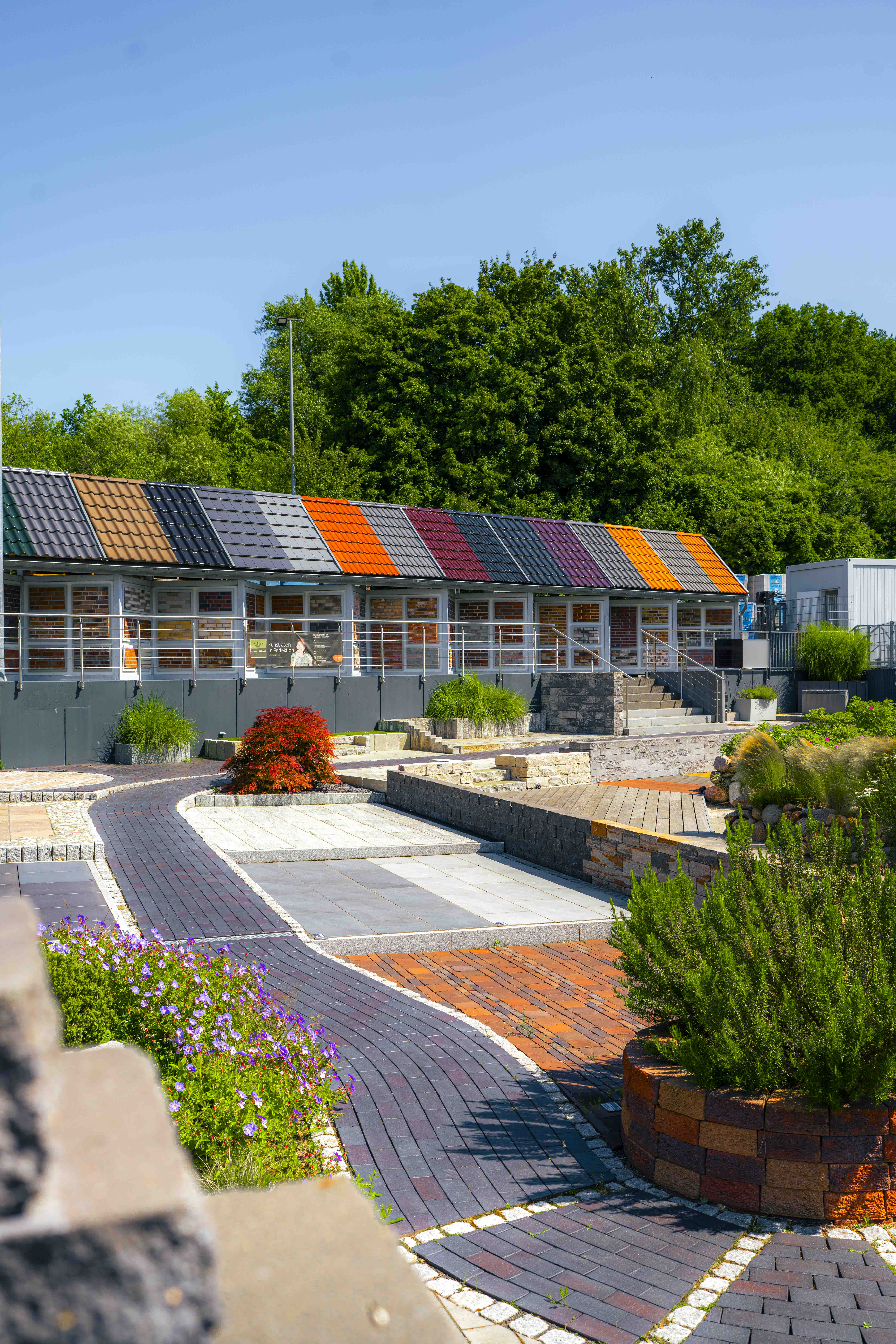 Mustergarten im Bauwelt Hummelsbüttel mit Pflaster- und Terrassenmustern sowie Dachziegel-Ausstellung in unterschiedlichen Farben vor grünem Baumbestand.